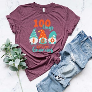 May include: Burgundy t-shirt with the text "100 Days with my Gnomies" and three gnome illustrations. The gnomes have colorful hats and the numbers 1, 0, and 0 on their bodies. Blue jeans are folded next to the shirt.