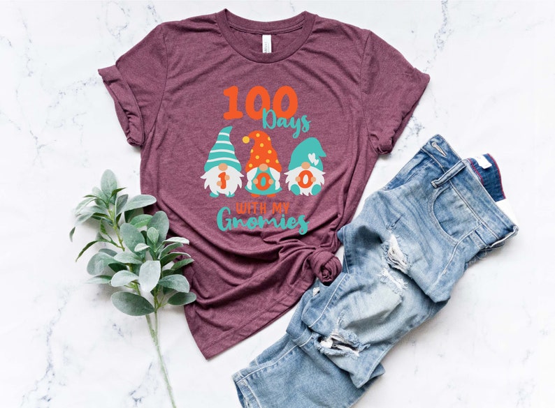 May include: Burgundy t-shirt with the text "100 Days with my Gnomies" and three gnome illustrations. The gnomes have colorful hats and the numbers 1, 0, and 0 on their bodies. Blue jeans are folded next to the shirt.