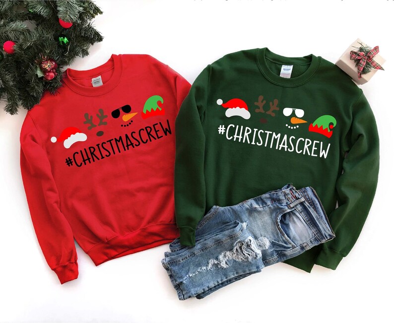 Pu&ograve; includere: Due felpe a tema natalizio, una rossa e una verde, con il testo "#CHRISTMASCREW" e una grafica di un pupazzo di neve, una renna e un elfo.