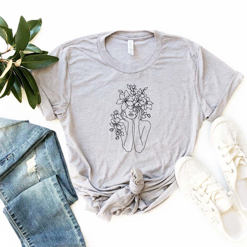 May include: A gray t-shirt with a black line drawing of a person with flowers covering their head and hands resting on their face.