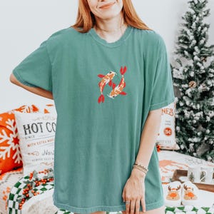 May include: A teal-colored t-shirt featuring an embroidered design of two koi fish. The fish are orange and white with red fins and tails. The shirt has a crew neck and short sleeves. The shirt is made of a soft, comfortable material.