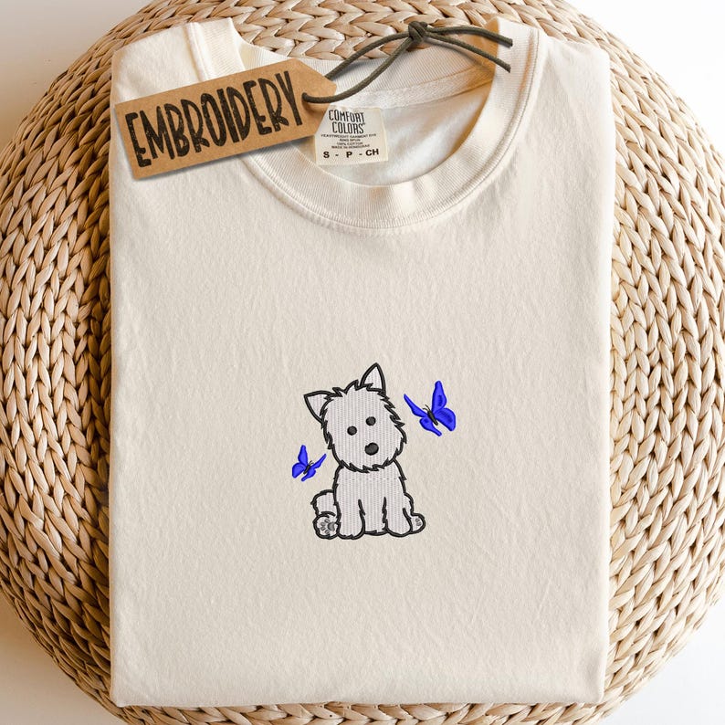 May include: Cream-colored t-shirt with an embroidered cartoon dog and two blue butterflies. The shirt has a tag that says "EMBROIDERY". The shirt is folded neatly.