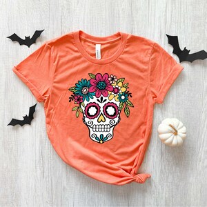 May include: Orange t-shirt with a white sugar skull design. The skull is decorated with colorful flowers and has black eyes and a black mouth.