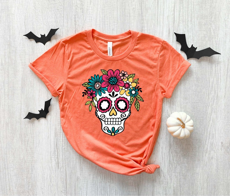 May include: Orange t-shirt with a white sugar skull design. The skull is decorated with colorful flowers and has black eyes and a black mouth.
