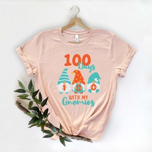 May include: A light pink t-shirt with the text "100 Days with my Gnomies" and three gnome illustrations. The gnomes have colorful hats and the number "1", "0", and "0" on their bodies. The shirt is hanging on a wooden hanger.