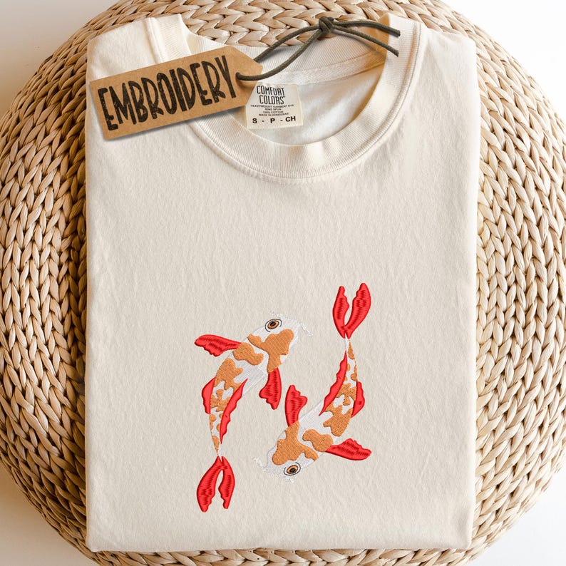 May include: Cream-colored t-shirt with embroidered koi fish design. The shirt features two koi fish in orange, red, and white. The tag reads "EMBROIDERY". The shirt is folded on a woven surface.