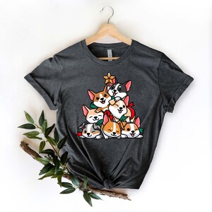 Puede incluir: Una camiseta gris oscuro con un dise&ntilde;o de &aacute;rbol de Navidad con una pila de corgis de dibujos animados. Los corgis llevan accesorios navide&ntilde;os rojos y verdes.