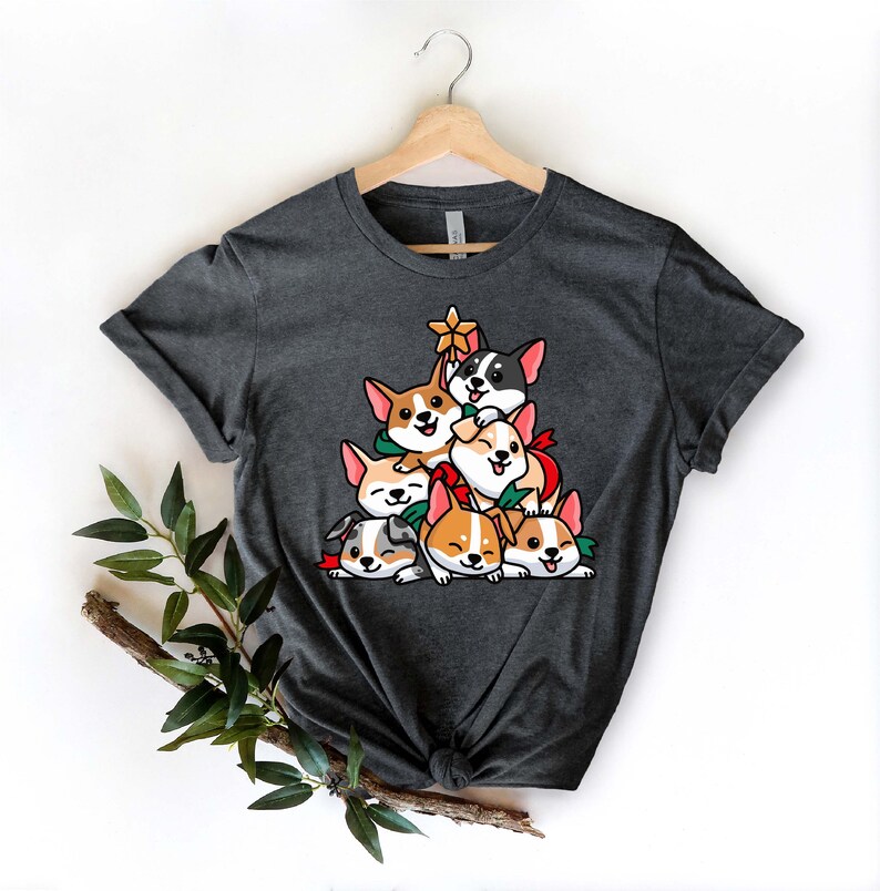 Puede incluir: Una camiseta gris oscuro con un dise&ntilde;o de &aacute;rbol de Navidad con una pila de corgis de dibujos animados. Los corgis llevan accesorios navide&ntilde;os rojos y verdes.