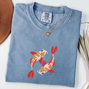 May include: A light blue Comfort Colors t-shirt featuring an embroidered design of two koi fish. The fish are orange, white, and red. A gold necklace with a small pendant rests on the shirt. The shirt is size small.