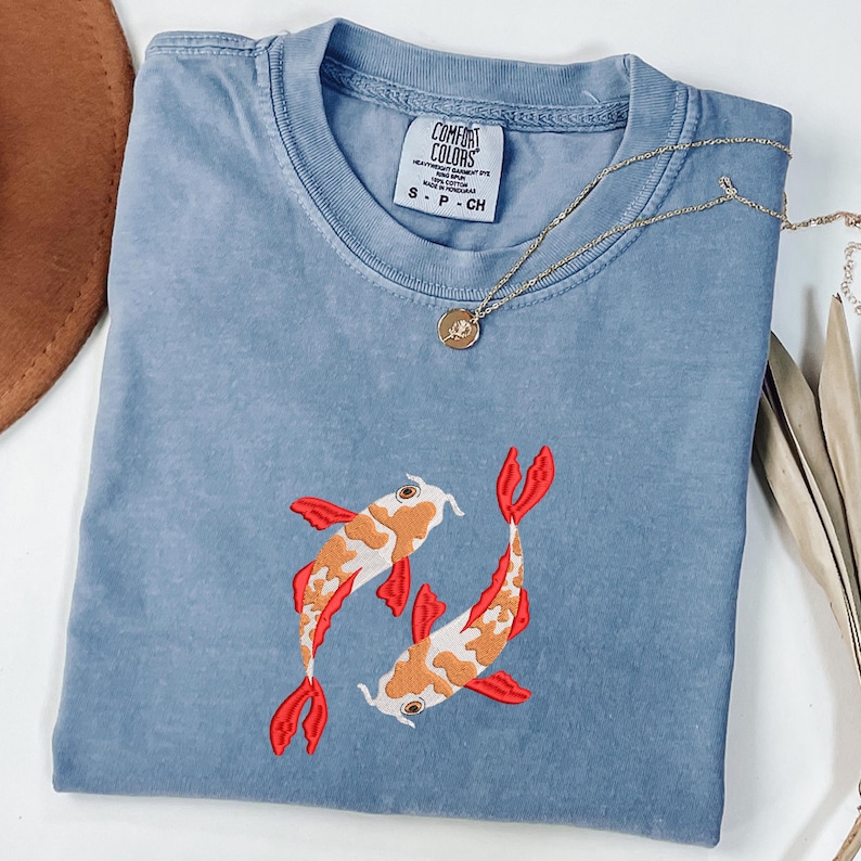 May include: A light blue Comfort Colors t-shirt featuring an embroidered design of two koi fish. The fish are orange, white, and red. A gold necklace with a small pendant rests on the shirt. The shirt is size small.