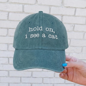 May include: Teal baseball cap with the embroidered text "hold on, i see a cat" in white. The cap has a curved brim and a fabric strap closure. The hat is made of a soft, washed cotton material.