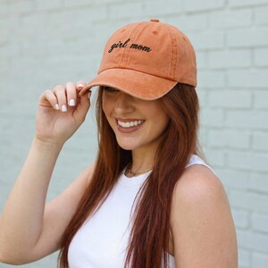 Puede incluir: Gorra de b&eacute;isbol naranja con las palabras "girl mom" bordadas en escritura negra. La gorra la lleva una persona con pelo largo rojo y una camiseta blanca. La gorra tiene una visera curva.