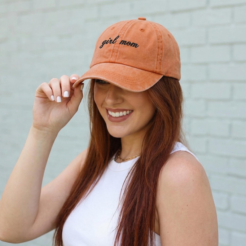 Puede incluir: Gorra de b&eacute;isbol naranja con las palabras "girl mom" bordadas en escritura negra. La gorra la lleva una persona con pelo largo rojo y una camiseta blanca. La gorra tiene una visera curva.