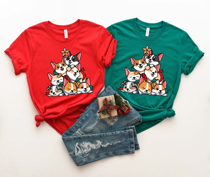 Puede incluir: Dos camisetas con un dise&ntilde;o de &aacute;rbol de Navidad con una pila de perros corgi. La camiseta roja tiene una estrella blanca en la parte superior del &aacute;rbol y la camiseta verde tiene una estrella amarilla en la parte superior del &aacute;rbol.
