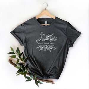 May include: A dark gray t-shirt with a white floral design and the text "YOUR NAME HERE" printed on the front.