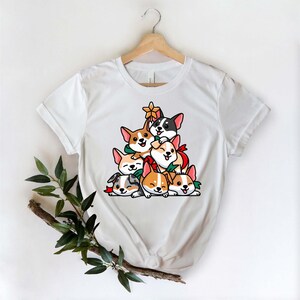 Puede incluir: Camiseta blanca con un dise&ntilde;o de &aacute;rbol de Navidad hecho de corgis de dibujos animados. Los corgis llevan lazos rojos y verdes y est&aacute;n apilados uno encima del otro.