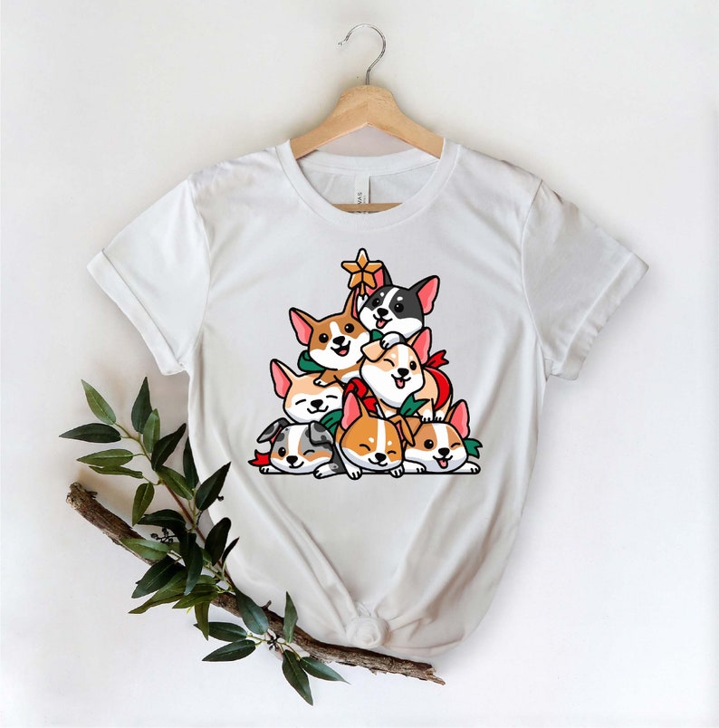 Puede incluir: Camiseta blanca con un dise&ntilde;o de &aacute;rbol de Navidad hecho de corgis de dibujos animados. Los corgis llevan lazos rojos y verdes y est&aacute;n apilados uno encima del otro.