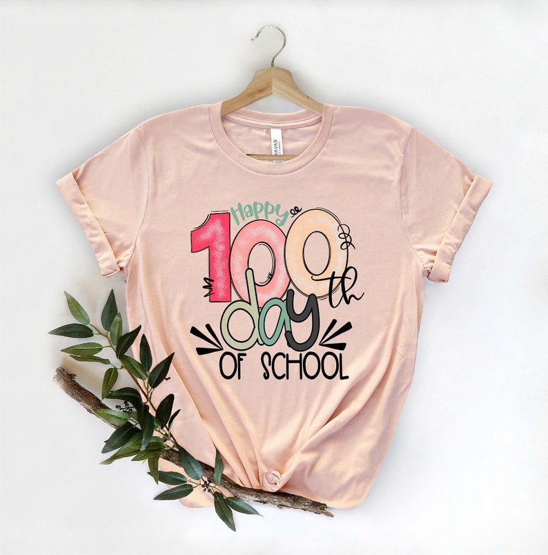 May include: A peach-colored t-shirt with a graphic design that says "Happy 100th day of school". The numbers 100 are in a colorful, layered style.