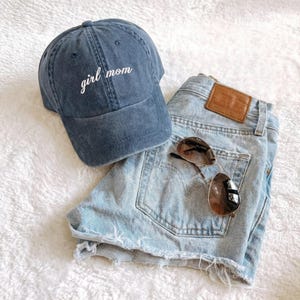 Puede incluir: Una gorra de b&eacute;isbol azul marino con el texto bordado "girl mom" se encuentra junto a unos pantalones cortos vaqueros azul claro y unas gafas de sol de aviador. Los pantalones cortos tienen un parche de cuero marr&oacute;n.