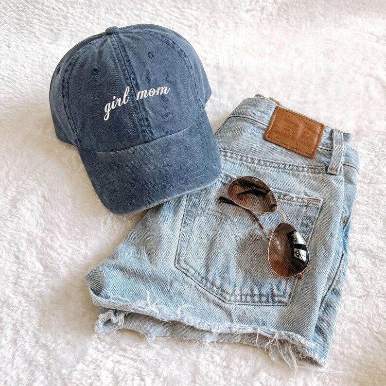 Puede incluir: Una gorra de b&eacute;isbol azul marino con el texto bordado "girl mom" se encuentra junto a unos pantalones cortos vaqueros azul claro y unas gafas de sol de aviador. Los pantalones cortos tienen un parche de cuero marr&oacute;n.