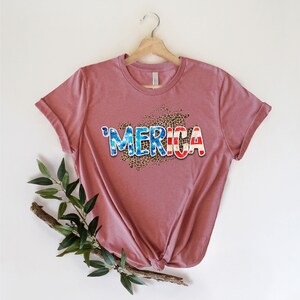 Op de afbeelding: Een mauve t-shirt met een grafische print van het woord "'MERICA" in blauw, wit en rood met een luipaardprint achtergrond.