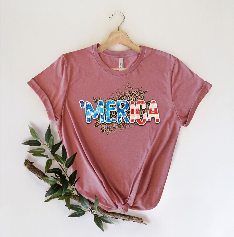 Op de afbeelding: Een mauve t-shirt met een grafische print van het woord "'MERICA" in blauw, wit en rood met een luipaardprint achtergrond.