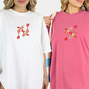 May include: White and pink t-shirts with a koi fish design. The koi fish are orange, white, and red. The t-shirts are oversized and have short sleeves. The white shirt is on the left, and the pink shirt is on the right.
