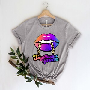 May include: Gray t-shirt with a rainbow-colored lips design with a gold chain. The text "Sagittarius Queen" is written below the lips.