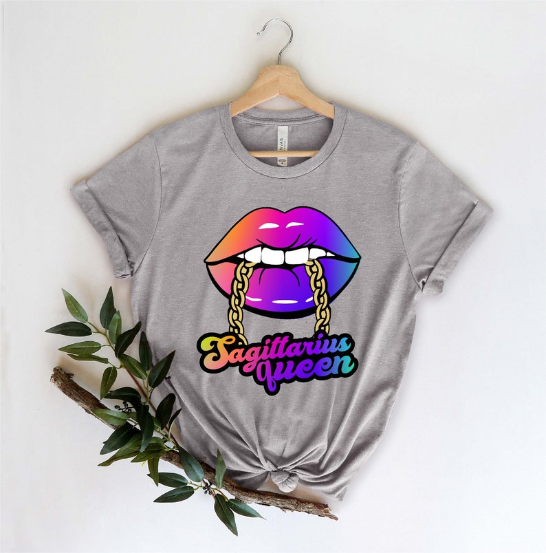 May include: Gray t-shirt with a rainbow-colored lips design with a gold chain. The text "Sagittarius Queen" is written below the lips.
