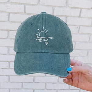 May include: Teal baseball cap with a white line drawing of a sun over water. The cap has a curved brim and an adjustable strap. The hat is made of a soft, washed cotton material.