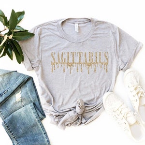 May include: A gray t-shirt with a gold glitter design that says "Sagittarius" with the letters dripping down.