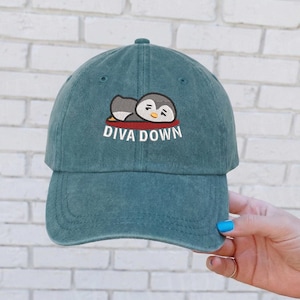 May include: Teal baseball cap with an embroidered design of a sleeping penguin and the words "DIVA DOWN" in white. The cap has a curved brim and a classic six-panel design, perfect for casual wear.