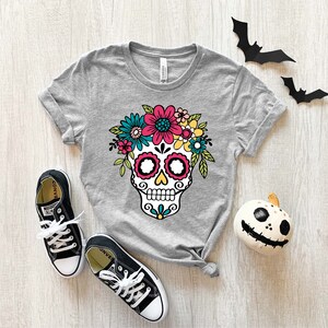 May include: Gray t-shirt with a colorful sugar skull design with flowers. The skull has black eyes, a white nose, and a pink mouth. The flowers are pink, yellow, blue, and green.