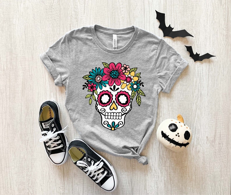 May include: Gray t-shirt with a colorful sugar skull design with flowers. The skull has black eyes, a white nose, and a pink mouth. The flowers are pink, yellow, blue, and green.