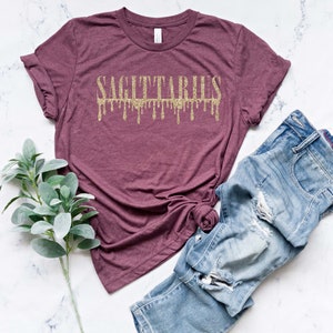 May include: A maroon t-shirt with the word "SAGITTARIUS" in gold glitter lettering. The letters are dripping down, creating a unique and stylish design.