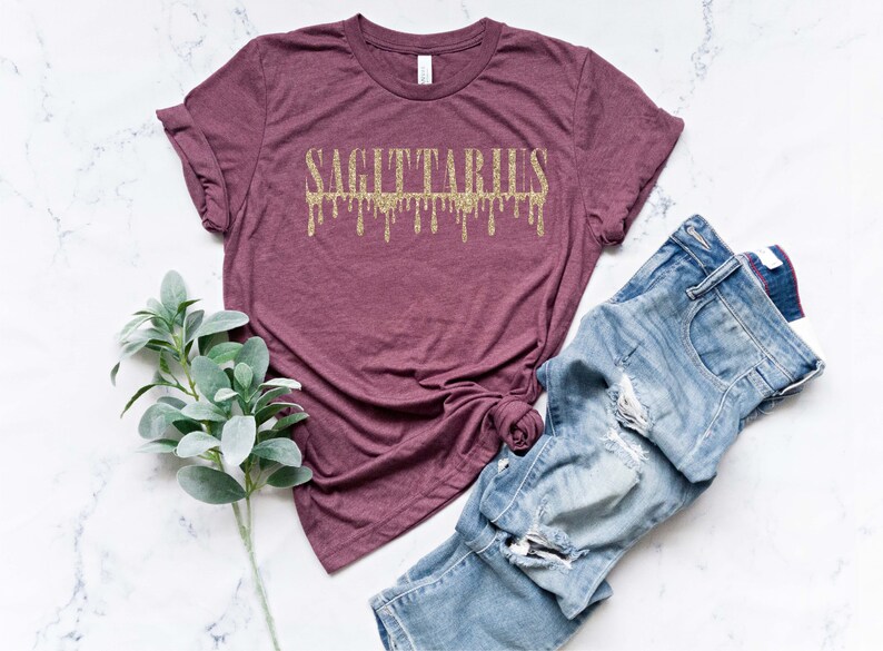 May include: A maroon t-shirt with the word "SAGITTARIUS" in gold glitter lettering. The letters are dripping down, creating a unique and stylish design.
