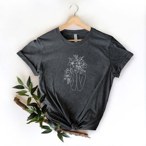 May include: A dark heather grey t-shirt with a white line drawing of a person with flowers in their hair.
