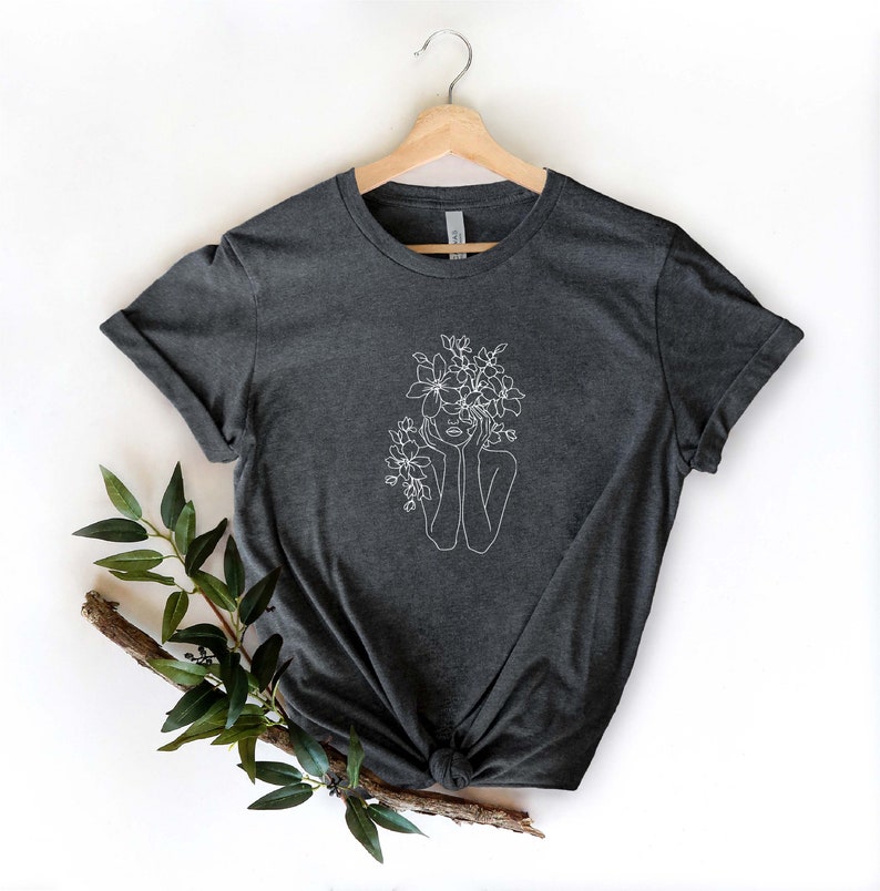 May include: A dark heather grey t-shirt with a white line drawing of a person with flowers in their hair.
