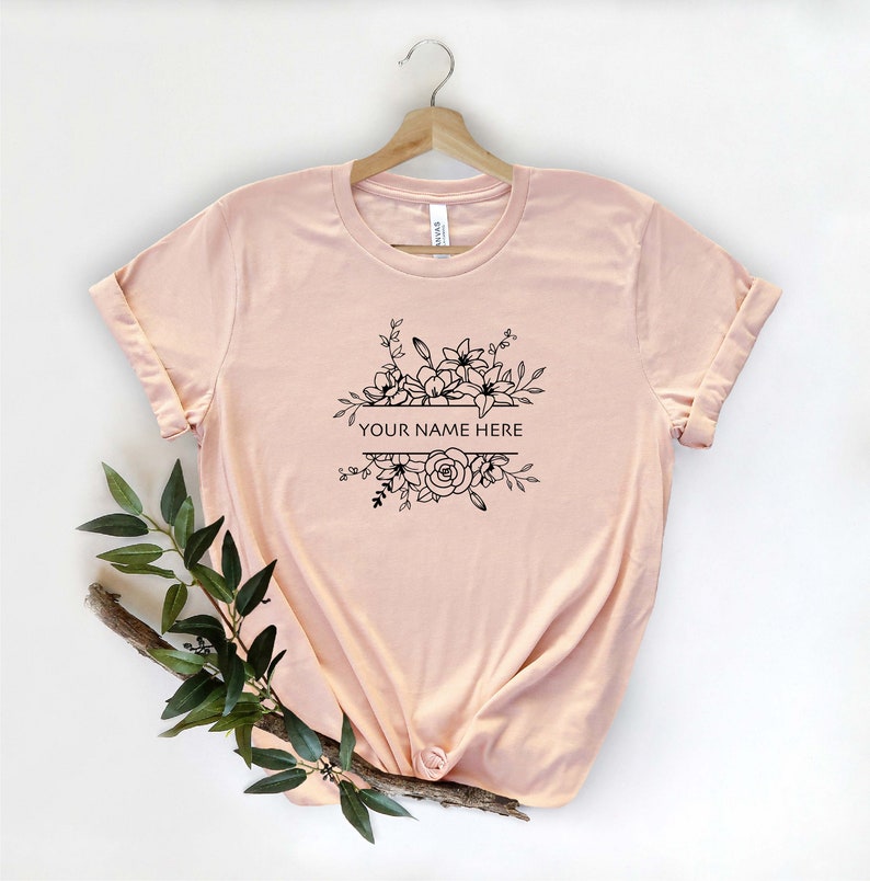 May include: A pale pink t-shirt with a black floral design and the text "YOUR NAME HERE" printed on the front.