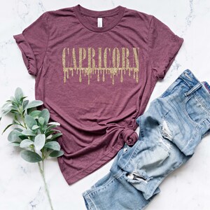 Pu&ograve; includere: Una maglietta color marsala con un designo di glitter dorato che dice "CAPRICORN" con gocce che cadono dalle lettere.