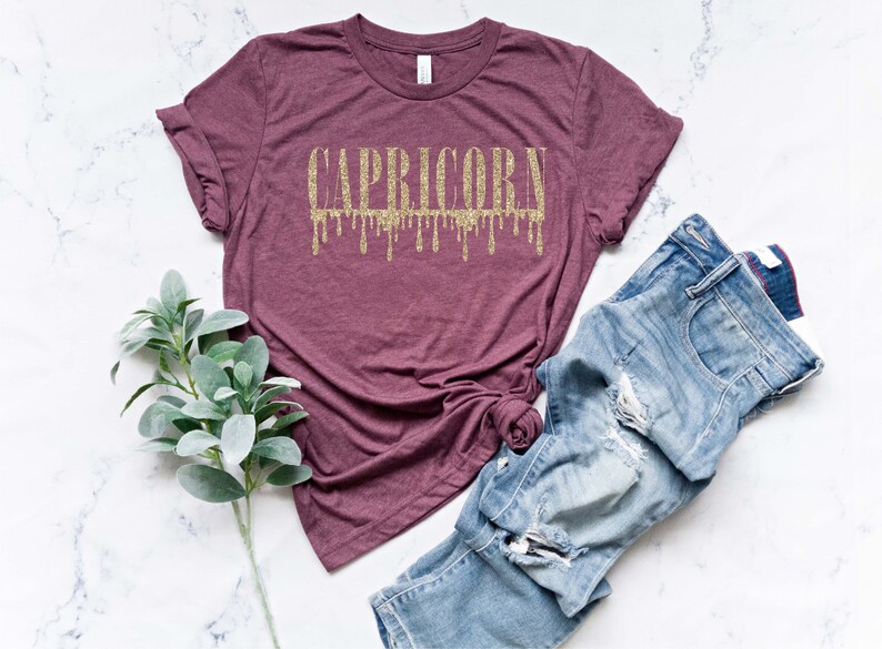 Pu&ograve; includere: Una maglietta color marsala con un designo di glitter dorato che dice "CAPRICORN" con gocce che cadono dalle lettere.