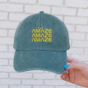May include: A teal baseball cap with the word "AMAZE" embroidered in yellow. The word is stacked three times with a stylized design above and below. The cap has a curved brim and a classic six-panel design.