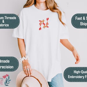 May include: White t-shirt with an embroidered design of two koi fish in shades of red and gold. The shirt features short sleeves and a relaxed fit. The image also includes text that says "Premium Threads & Fabrics", "Handmade With Precision", and "High-Quality Embroidery Finishing".