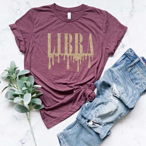 May include: A maroon t-shirt with a gold glitter "LIBRA" graphic with dripping accents. The shirt is laid flat on a white marble surface with a pair of blue jeans and a sprig of greenery.