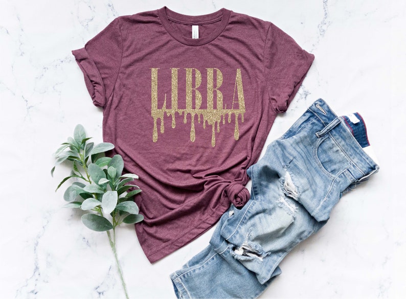 May include: A maroon t-shirt with a gold glitter "LIBRA" graphic with dripping accents. The shirt is laid flat on a white marble surface with a pair of blue jeans and a sprig of greenery.