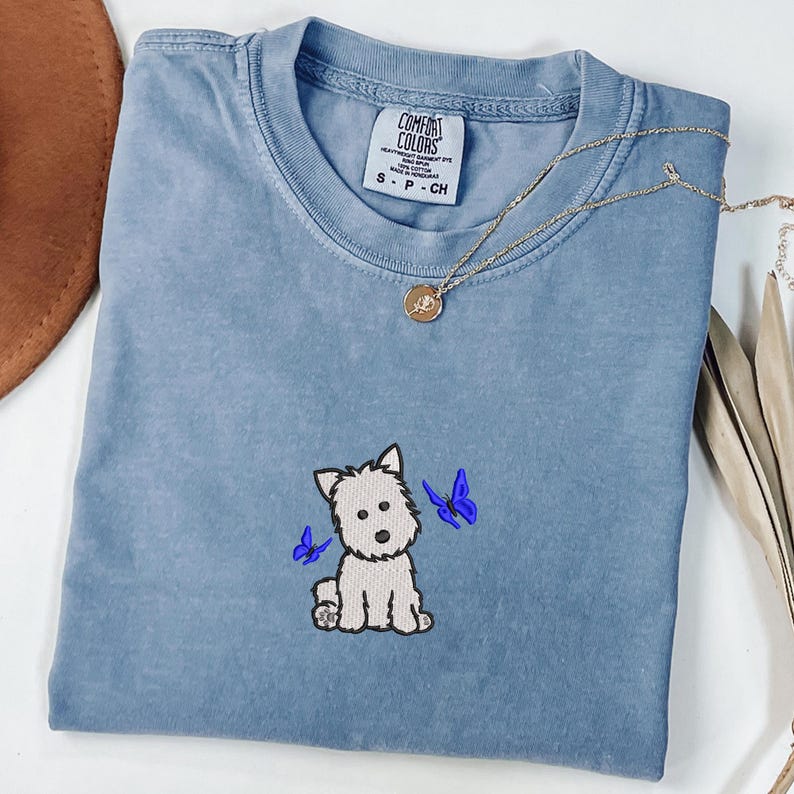 May include: A light blue Comfort Colors t-shirt featuring an embroidered white dog with two blue butterflies. A gold necklace with a pendant rests on the shirt. The shirt is size S-P-CH.