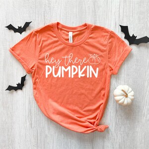 Puede incluir: Camiseta naranja de manga corta con el texto "hey there PUMPKIN" en blanco con un peque&ntilde;o gr&aacute;fico de calabaza.