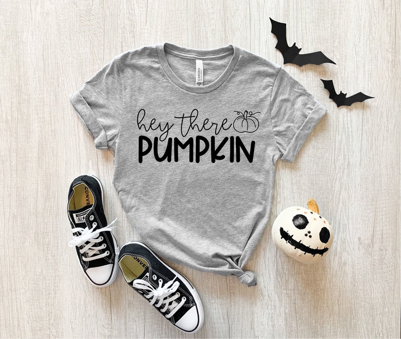 Puede incluir: Una camiseta gris con el texto "hey there pumpkin" en letras negras y un peque&ntilde;o gr&aacute;fico de calabaza. La camiseta est&aacute; colocada sobre una superficie de madera con un par de zapatillas negras y una calabaza blanca con una cara negra.