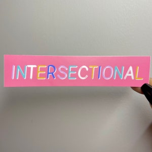May include: A pink rectangular sticker with the word "INTERSECTIONAL" in colorful, outlined letters. The sticker is held by a hand with a black painted fingernail against a gray background.