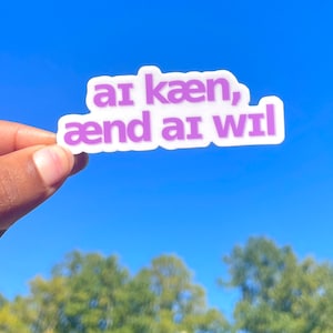 May include: A white sticker with the text "ar kæen, ænd ar wil" in purple letters. The sticker is held up against a bright blue sky with green trees in the background.